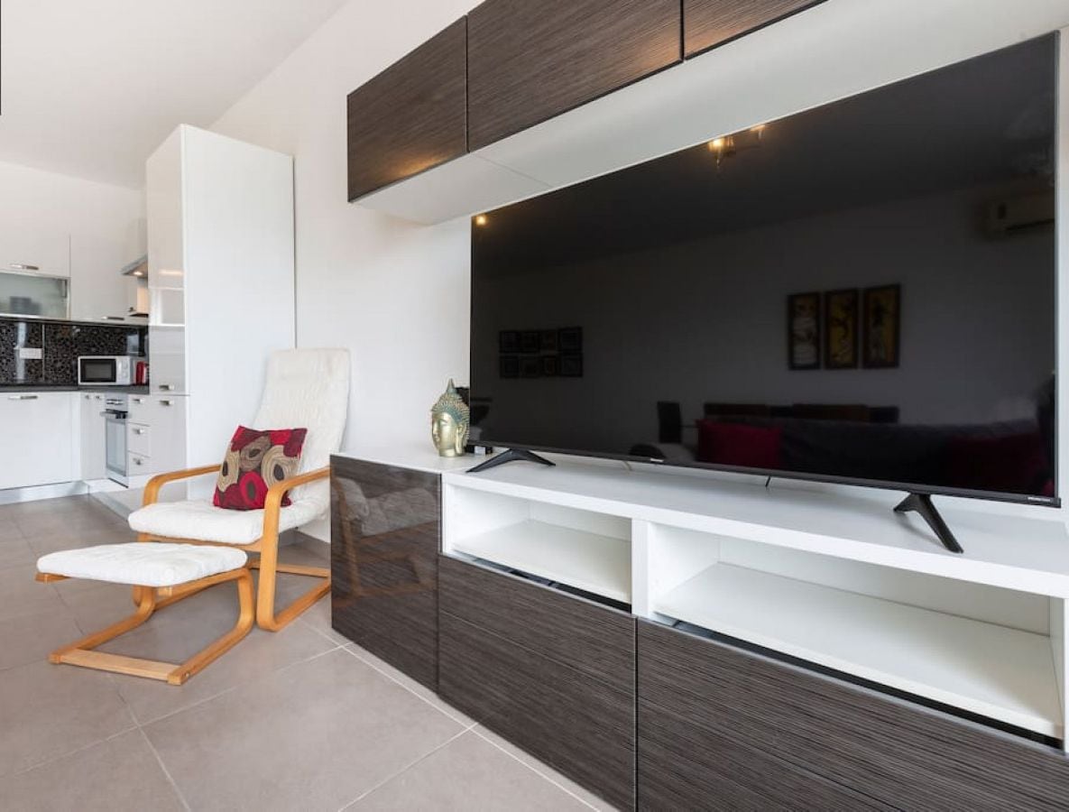 Un soggiorno moderno con un'ampia TV a schermo piatto, un divano bianco e una sedia in legno.