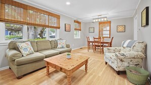 A spacious living room with a sofa, a coffee table, and a patterned armchair. There are large windows with blinds, a dining table with chairs, and a chandelier.