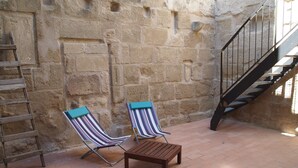 A stone-walled courtyard with two striped lounge chairs and a small wooden table. A metal ladder and a black staircase are also visible.