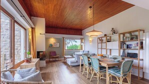A spacious room with a wooden ceiling, a dining area with wicker chairs, a bookshelf, and a view of the outdoors through large windows. - Maison Lamina - Welkeys (Anglet)