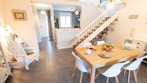 A dining area with a wooden table, white chairs, and a fruit bowl. A staircase with a white railing leads to another room.