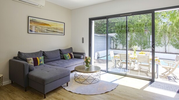 A modern living room with a grey sofa, a round coffee table, and a large sliding glass door leading to an outdoor patio area with a pool and furniture. - Villa Miarritze - Welkeys (Biarritz)