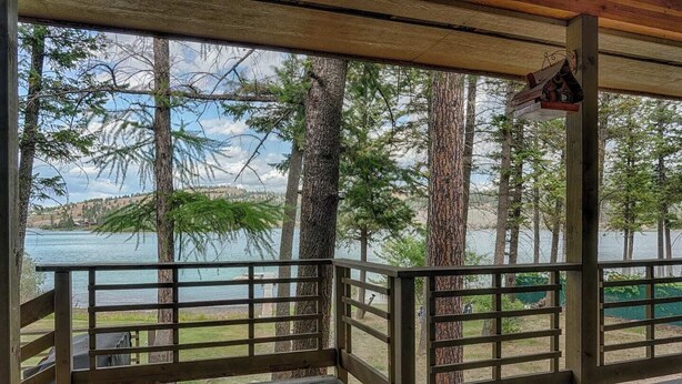 A wooden deck with a railing overlooking a lake, surrounded by tall trees. - Love on Lilac Lake, Private Dock and Lake Access (Kalispell)