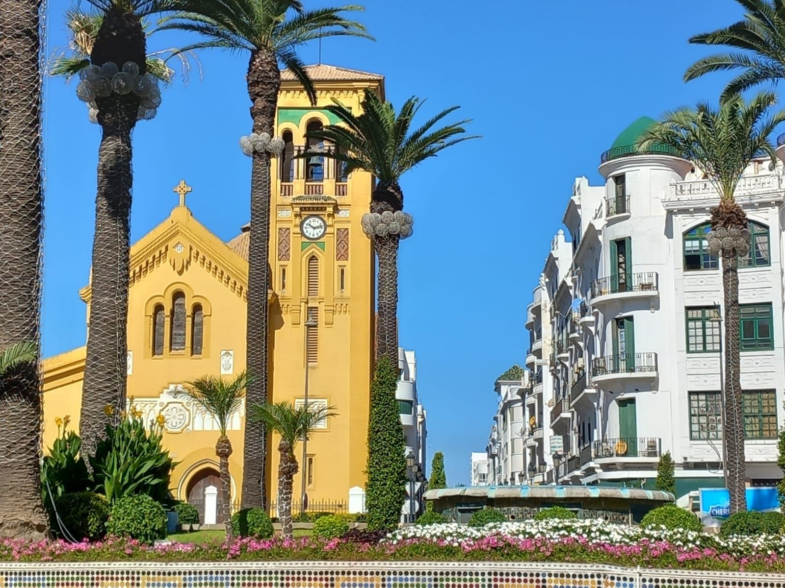 A yellow church with a clock tower, surrounded by palm trees and a flower garden.