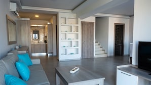 A modern living room with a sofa, coffee table, and TV stand. There is a kitchen area with a staircase leading to another level. - Enali Villas-Villa Artemi (Κίσσαμος)