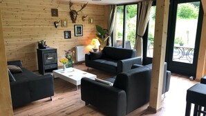 A cozy living room with a fireplace, wooden walls, and a large window with a view of greenery. - Ferienhaus im Sauerland (Winterberg)