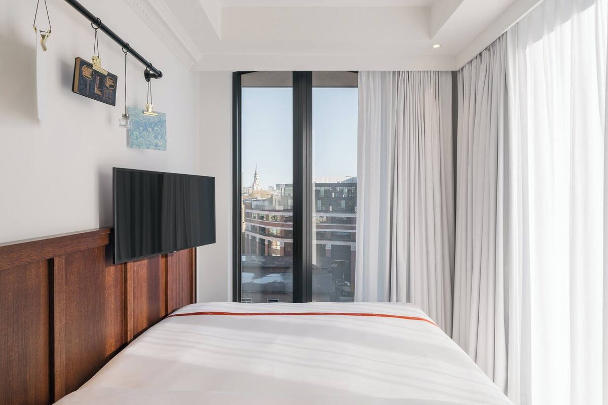 A hotel room with a large bed, a mounted TV, and a city view through a large window.