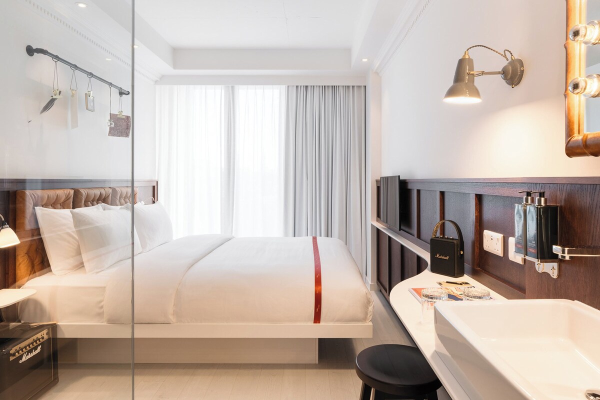 A modern hotel room with a bed, a vanity with a mirror, and a large window with curtains.