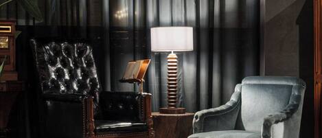 A room with a dark curtain backdrop, a black leather armchair, a blue velvet armchair, a wooden side table with a lamp, and a framed picture.