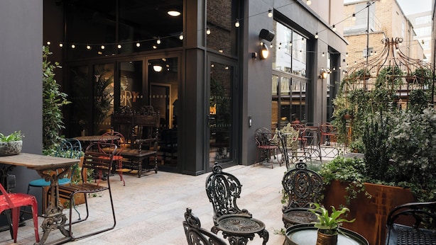 An outdoor cafe with tables and chairs, a brick building in the background, and string lights. - Ruby Stella Hotel London by IHG (London)