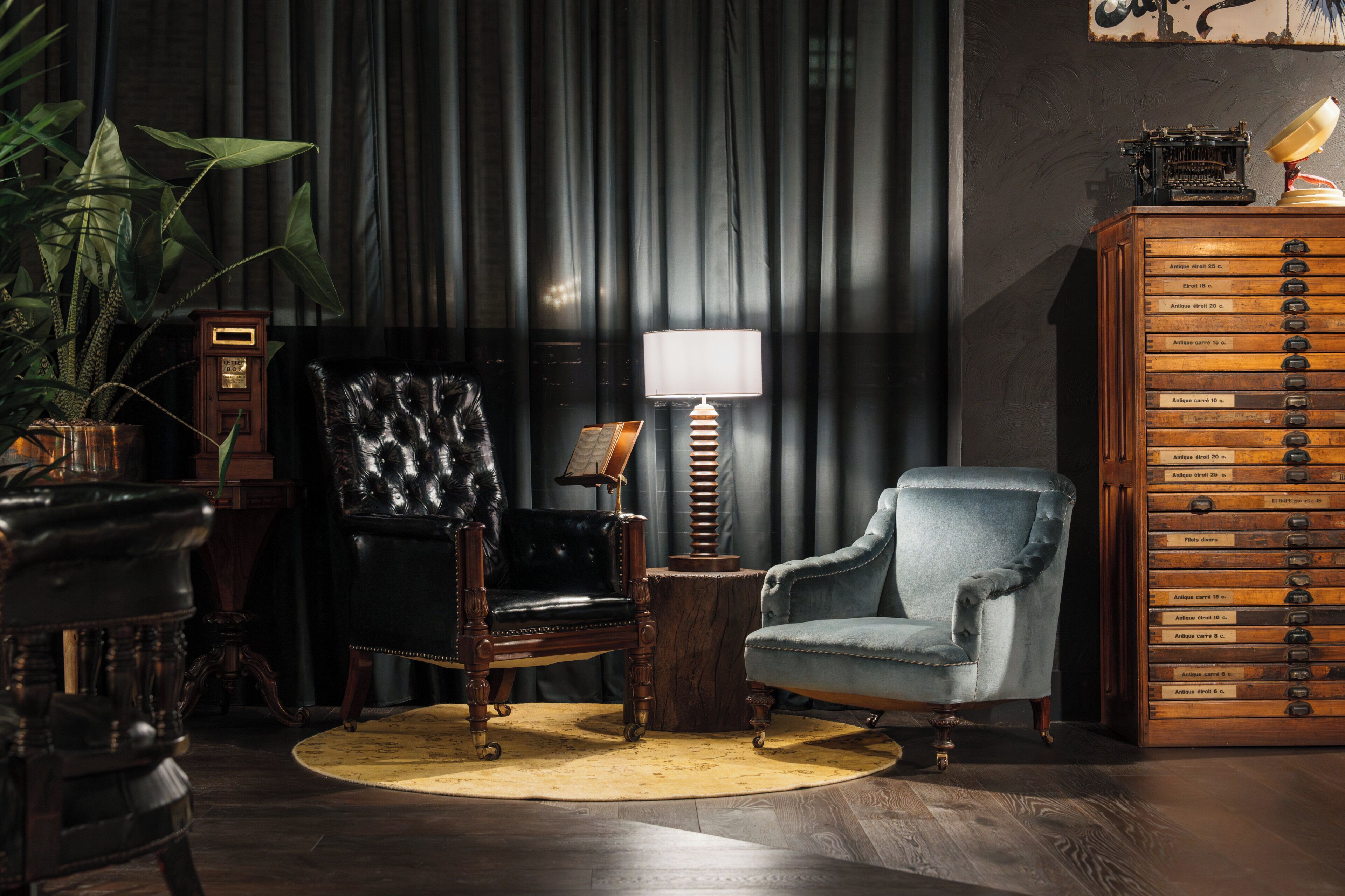 A room with a dark curtain, a desk with a lamp, a chair, a plant, and a wooden cabinet with drawers.