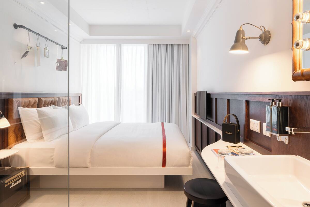 A modern hotel room with a bed, a vanity with a mirror, and a large window with curtains.