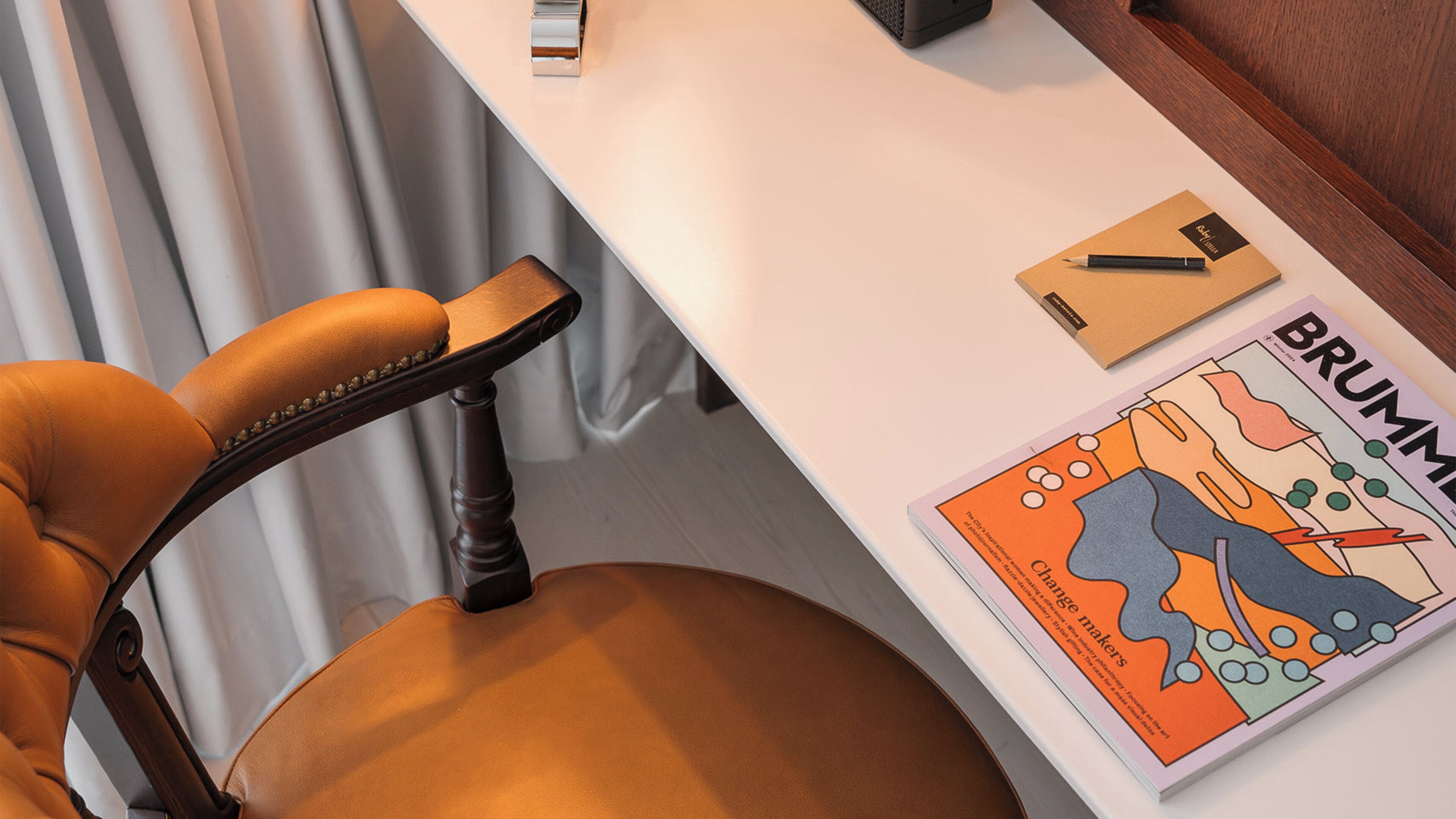 Una silla de oficina moderna con asiento de cuero y reposabrazos de madera, un escritorio blanco con una revista y un bolígrafo, y el borde de un escritorio de madera.