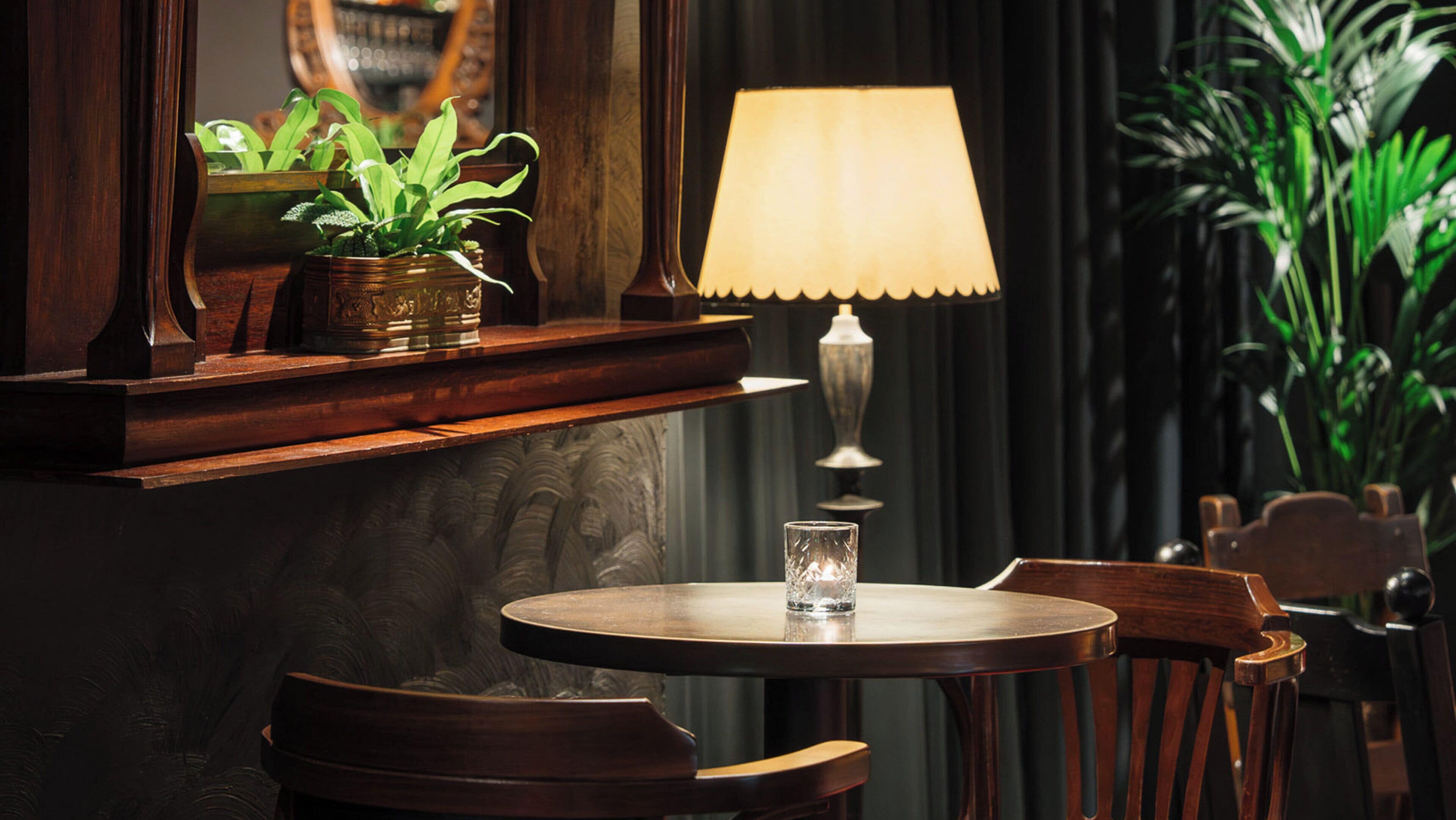 A round wooden table with a glass, a lamp, and a plant on a wooden shelf.