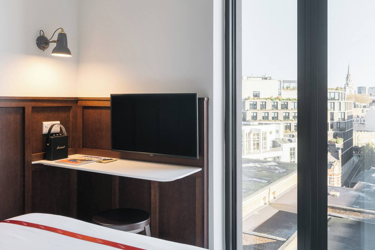A hotel room with a large window, a flat-screen TV, a Marshall speaker, a small stool, and a bed with a red stripe.