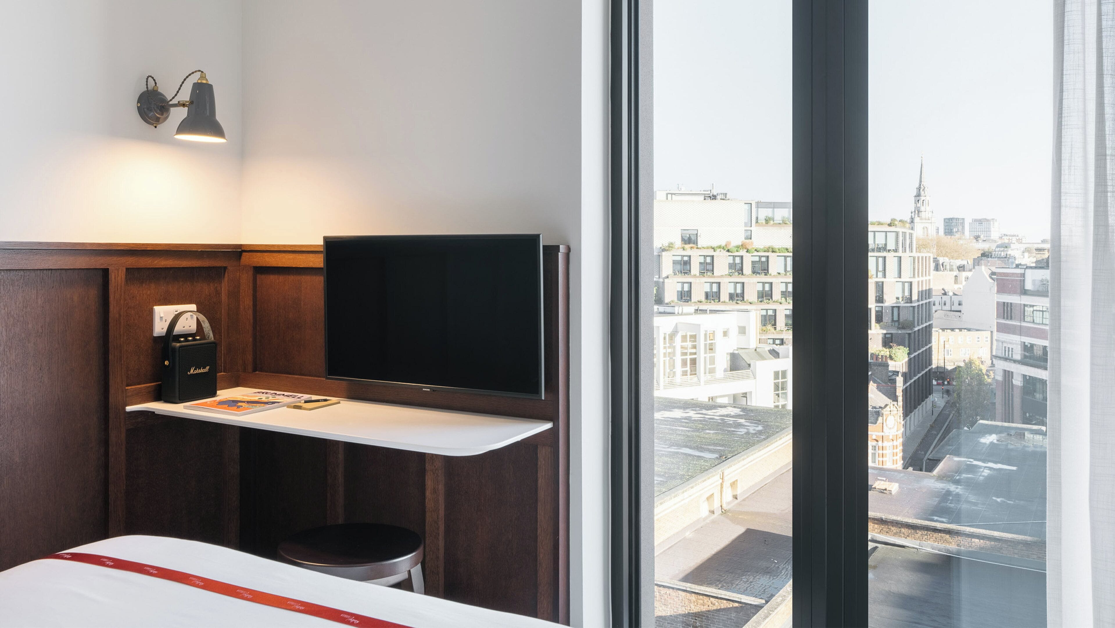 A hotel room with a large window, a flat-screen TV, a Marshall speaker, a small stool, and a bed with a red stripe.