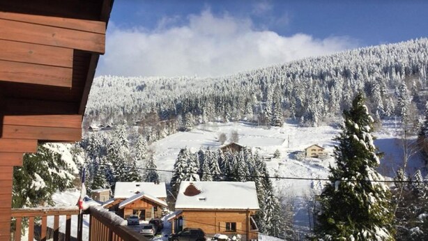 A wooden cabin with a snow-covered landscape, a clear sky, and a forested mountain in the background.