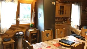 A cozy cabin interior with a wooden dining table, chairs, a refrigerator, and a window with curtains.