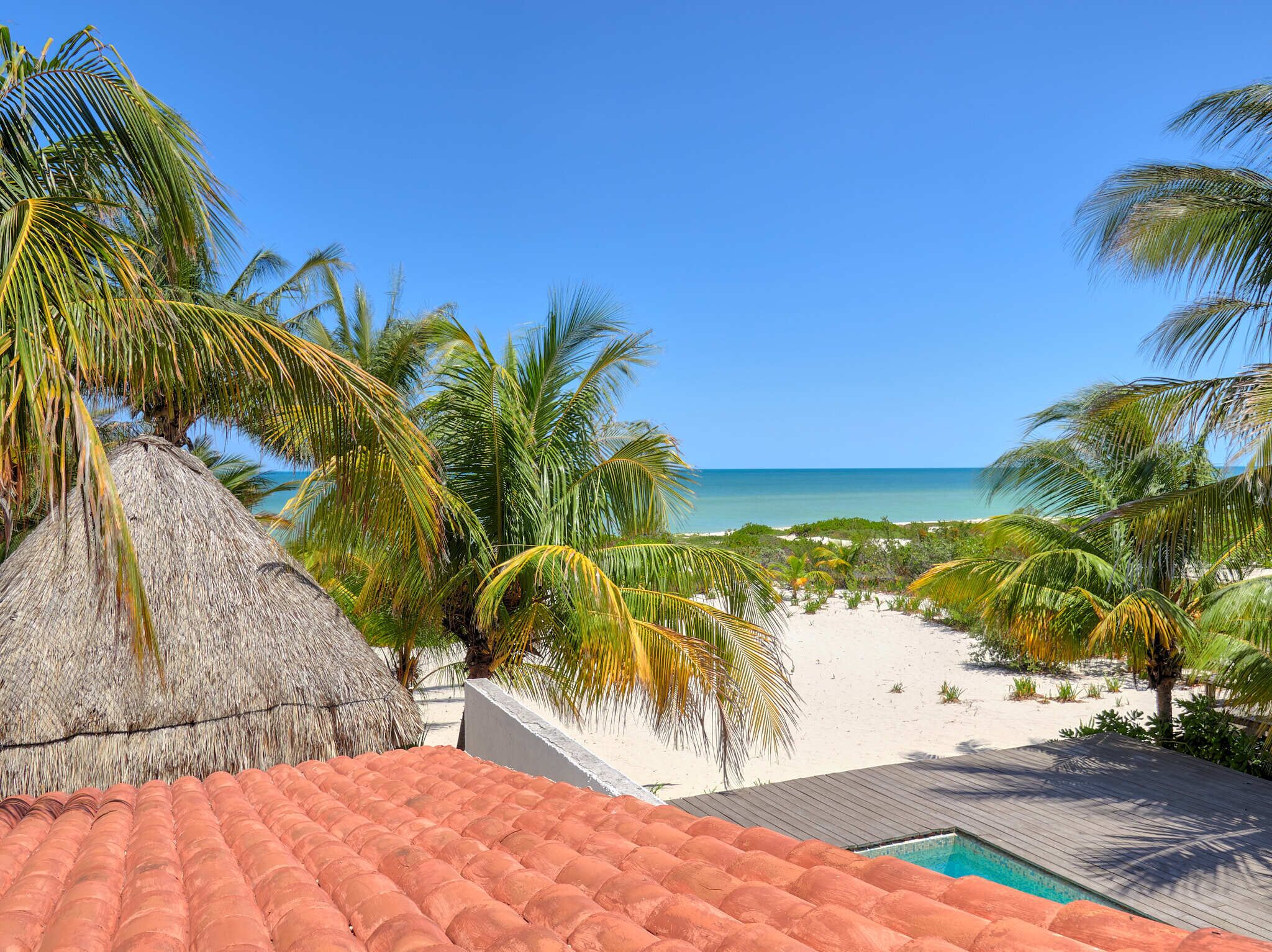 Blick auf einen Strand mit einer strohgedeckten Struktur, Palmen und blauem Himmel.
