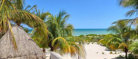 A beach view with a thatched roof structure, palm trees, and a clear blue sky.