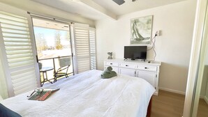 Un dormitorio con cama, televisor en una cómoda, ventana con persianas y vista al exterior.