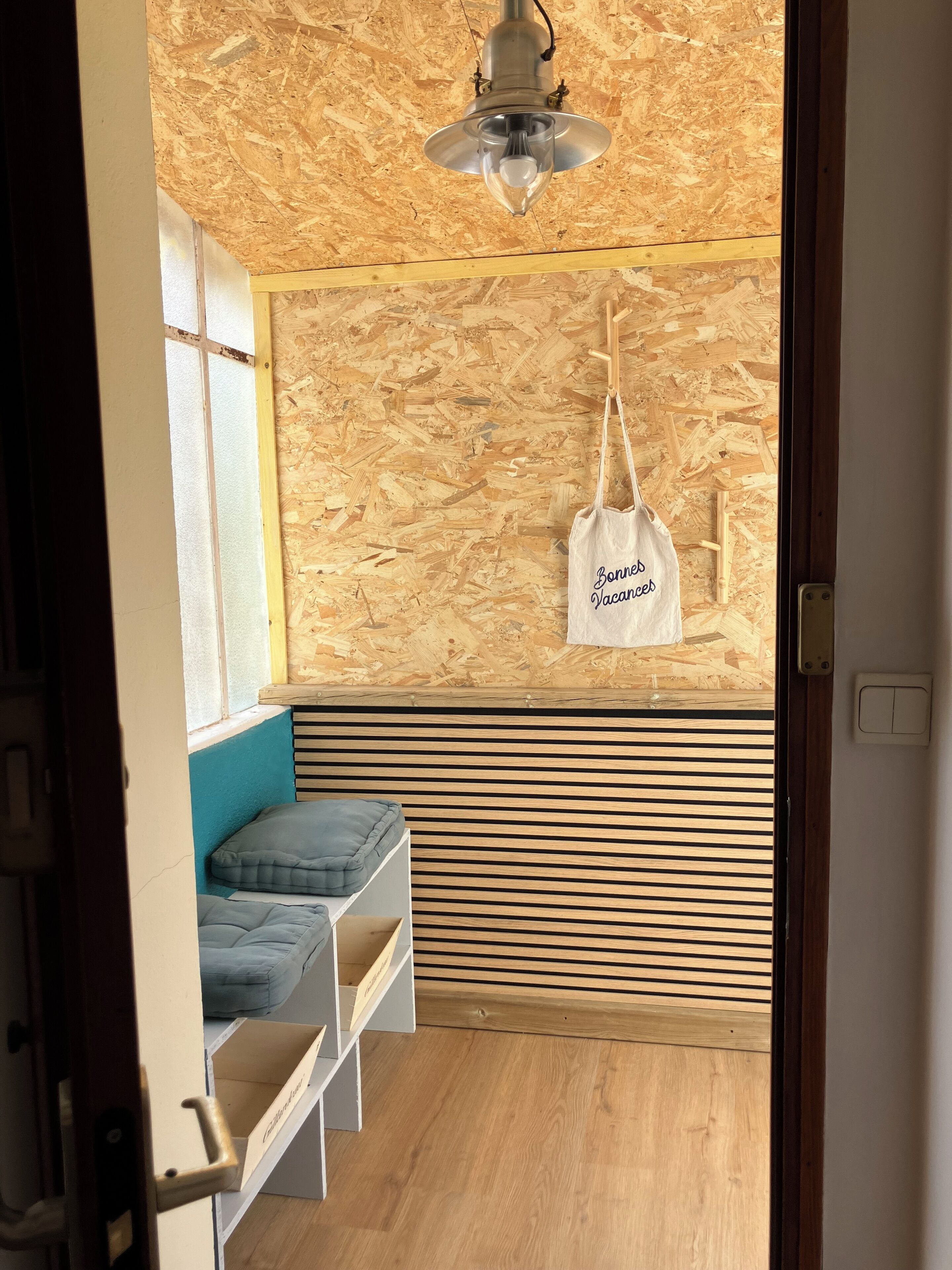 A room with a slatted wooden wall, a hanging light fixture, a bench with cushions, and a white bag with text.