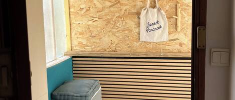 A room with a slatted wooden wall, a hanging light fixture, a bench with cushions, and a white bag with text.