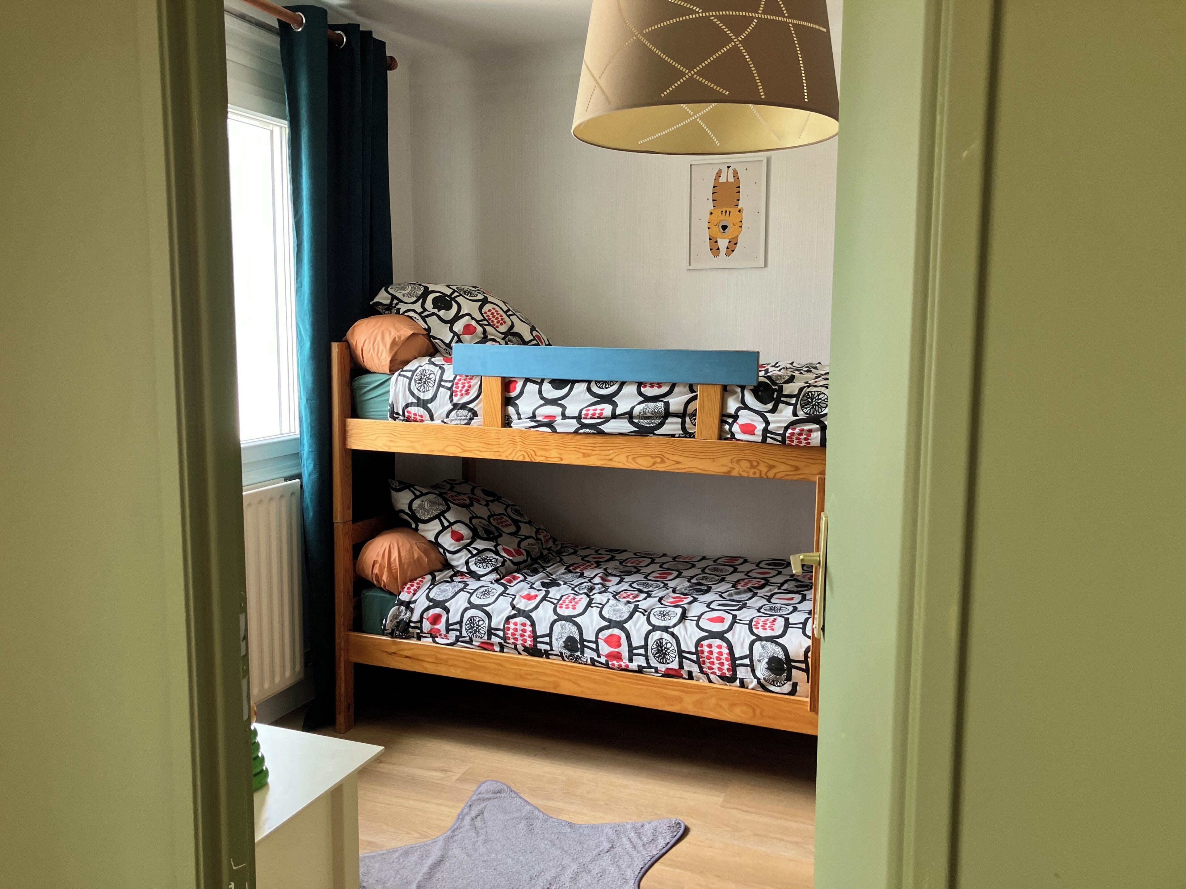 A bunk bed with patterned bedding, a window with curtains, a radiator, and a hanging lamp.