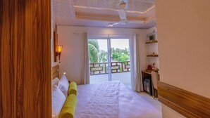 A hotel room with a large bed, a ceiling fan, a balcony with a view of greenery, and a small desk with a chair.
