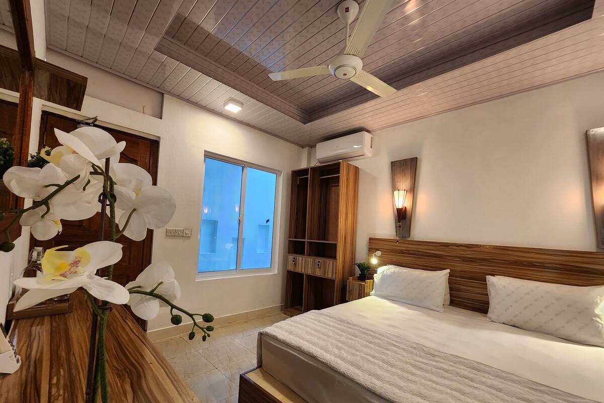 Un dormitorio con una cama grande, cabecera de madera y ventilador de techo. Se aprecia una ventana con vistas al exterior, y un armario de madera con estantes incorporados.