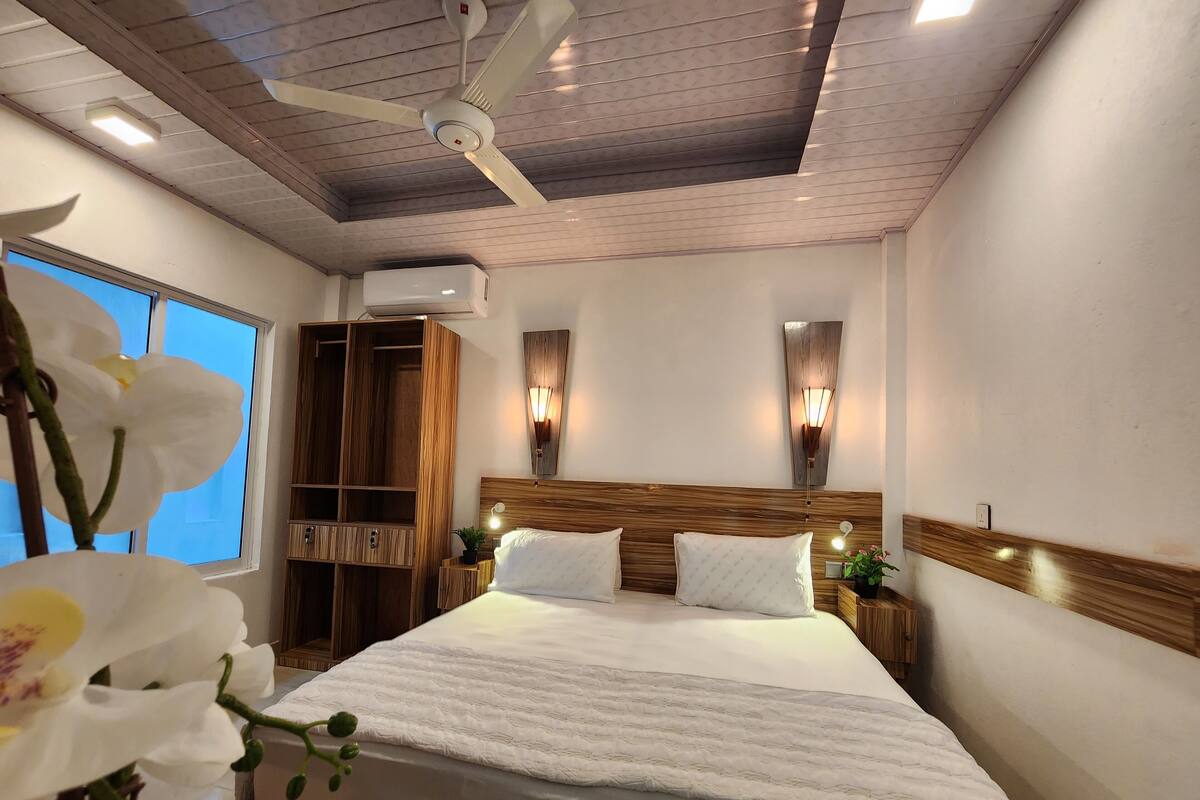 Una habitación de hotel con una cama grande, cabecero de madera, dos mesitas de noche con lámparas, un ventilador de techo y un equipo de aire acondicionado.