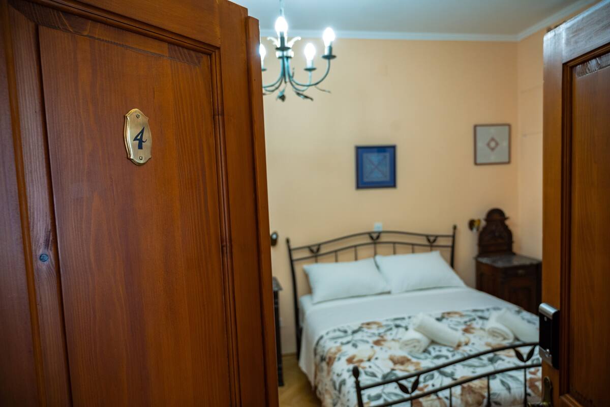 A bedroom with a wooden door marked with the number 4, a bed with a floral bedspread, a chandelier, and a framed picture on the wall.