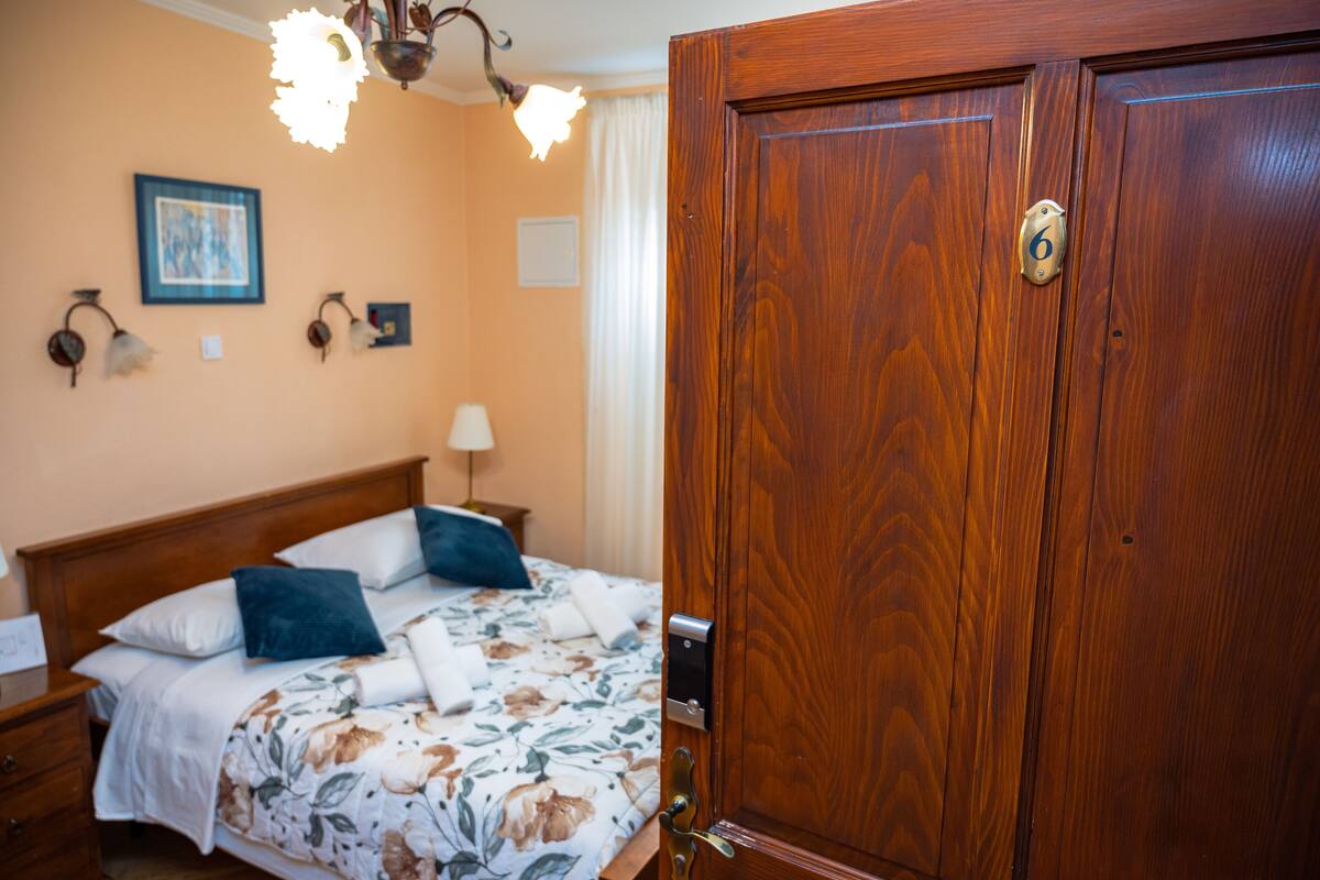 A hotel room with a wooden wardrobe, a bed with floral bedding, a nightstand, and a wall-mounted lamp.