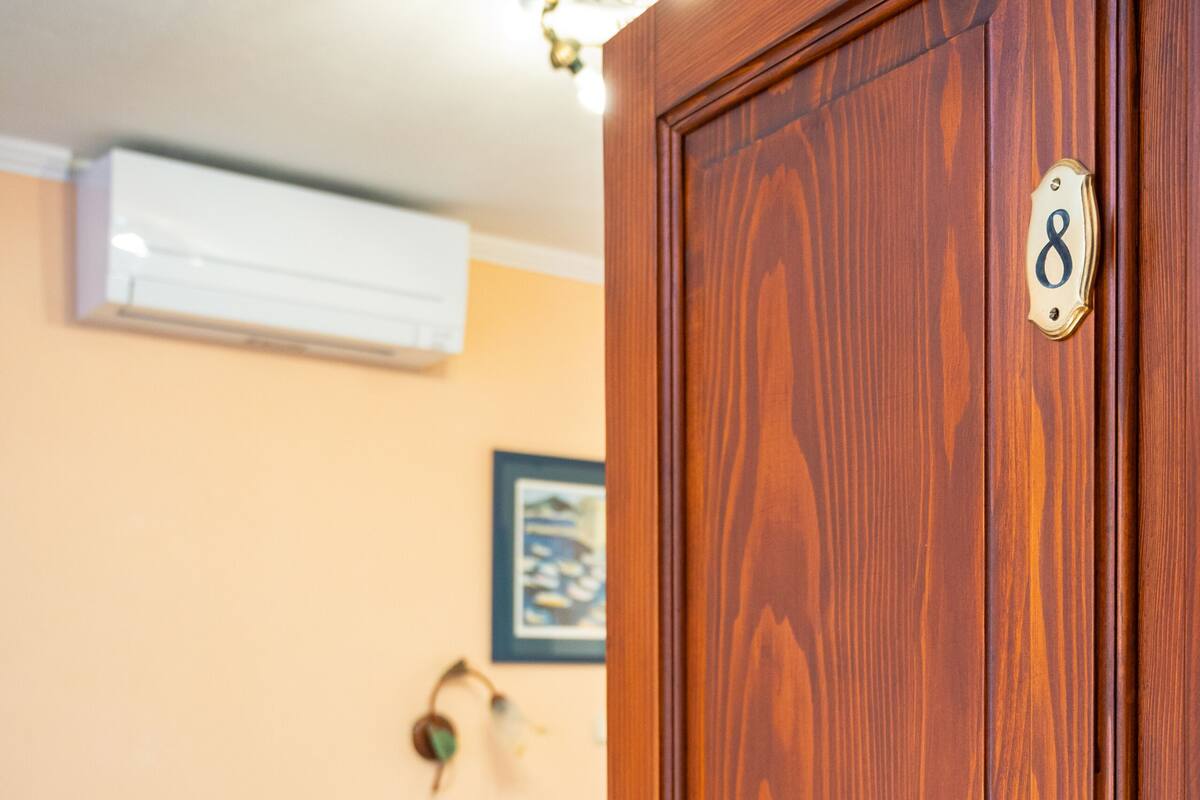 A wooden door with a brass handle and a number plate, a bed with a white sheet and a floral pattern, a bedside table with a lamp, a chair, and a wall-mounted air conditioner.