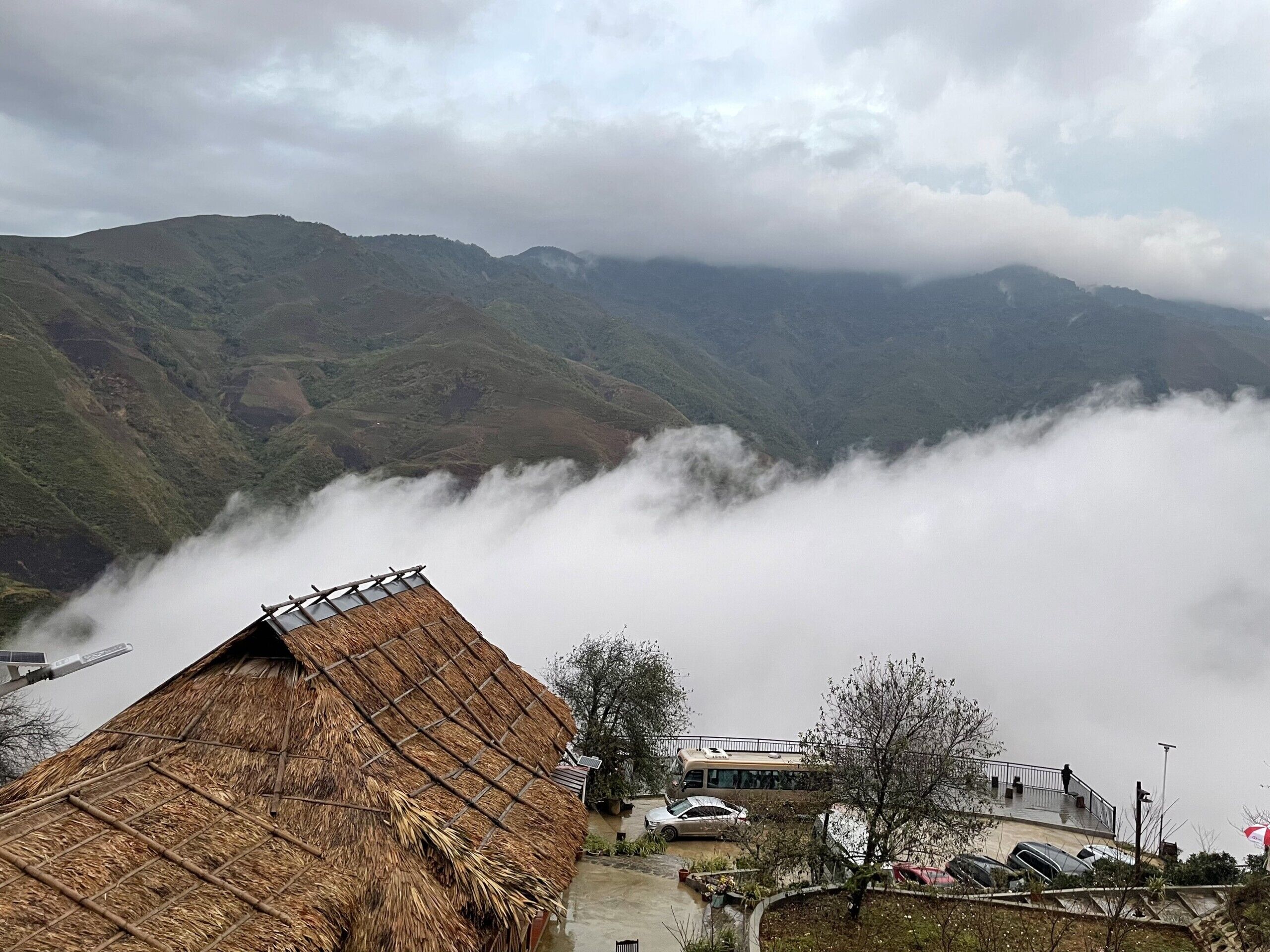 A mountain landscape with a thatched-roof building, a bus, and parked cars.