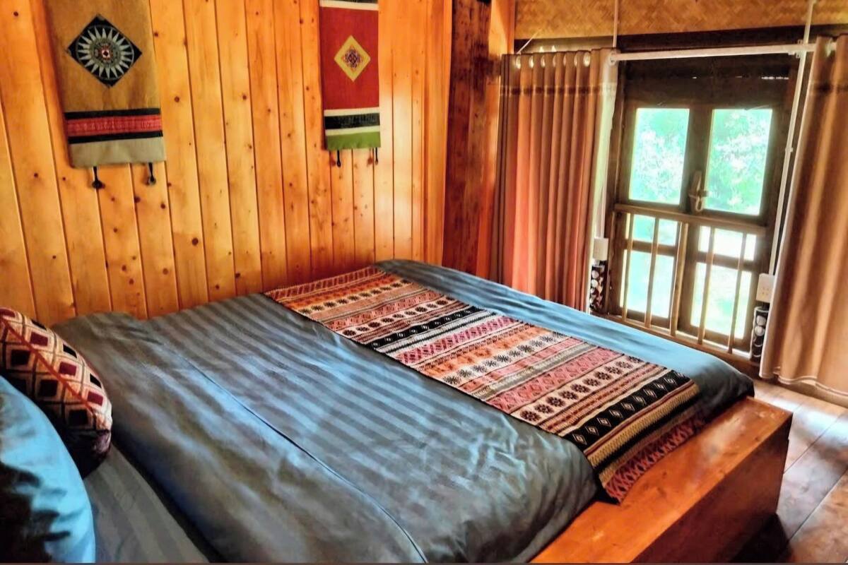 Um quarto de madeira com uma cama, um banco e uma janela com cortinas.