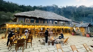 Un área de comedor al aire libre con mesas y sillas de madera, un edificio con techo de paja y un brasero.
