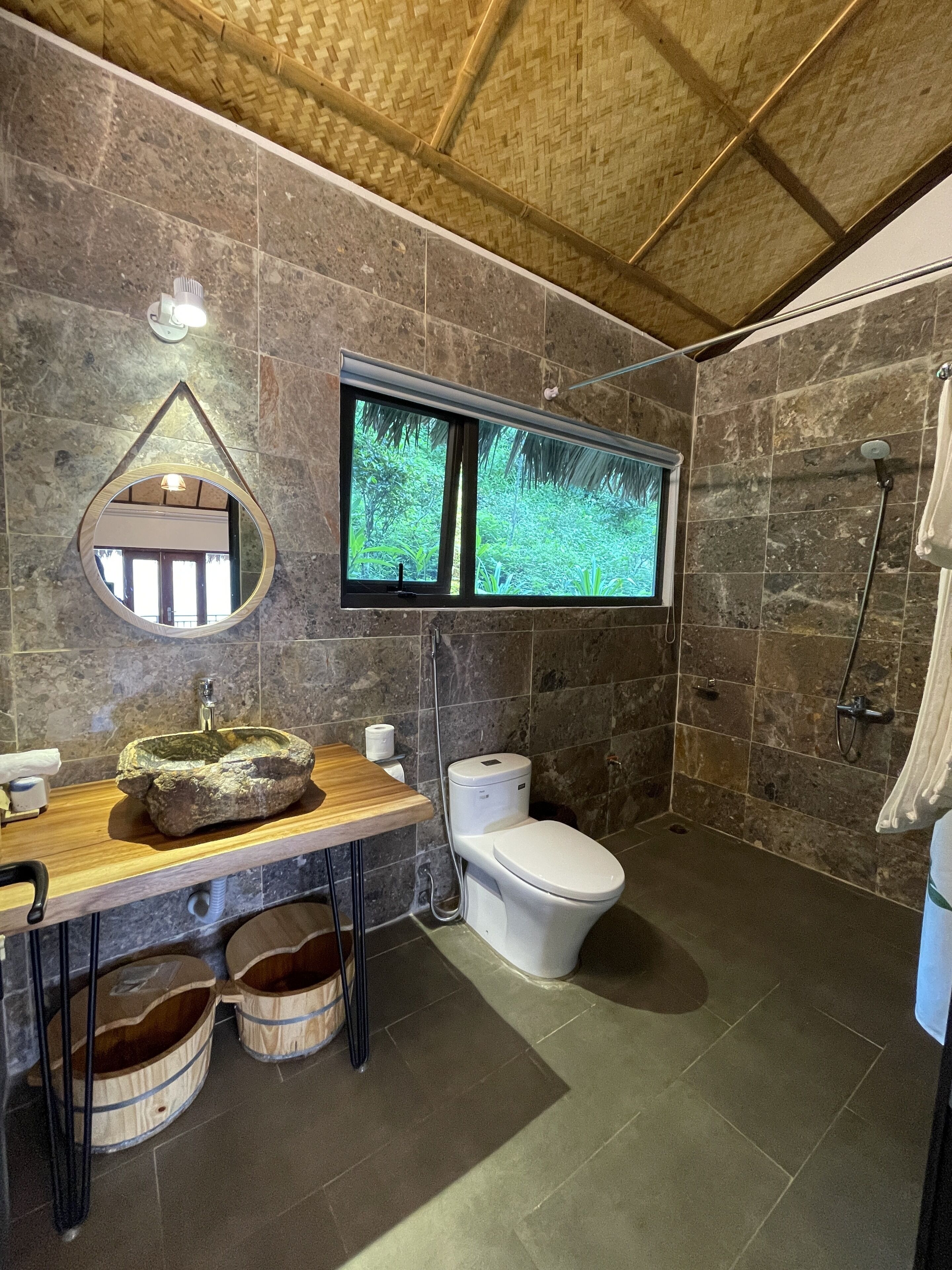 A bathroom with a stone wall, a wooden vanity, a round mirror, a toilet, and a shower area.