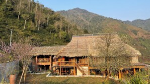 Una casa de madera tradicional con techo de paja, rodeada de exuberante vegetación y montañas al fondo.