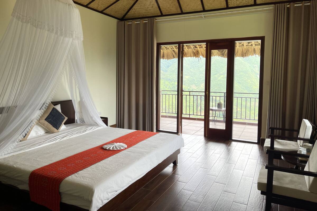 Quarto com cama, um objeto decorativo vermelho, varanda com vista para o verde e uma cadeira.
