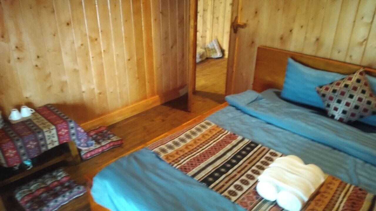 A wooden-paneled room with a bed, a bench with blankets, and a door leading to another room.