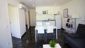 Une cuisine moderne avec des meubles blancs, un four à micro-ondes et un réfrigérateur. On y trouve un coin repas avec une table et des chaises blanches. Un canapé gris se trouve dans l’espace salon.
