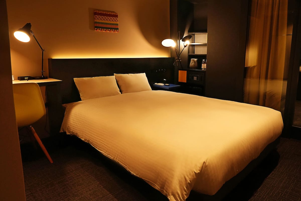A neatly made bed with a headboard, two bedside lamps, a small table, and a chair in a dimly lit room.