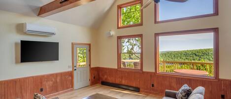 Un vaste salon doté d’un plafond en bois, de grandes fenêtres offrant une vue sur une forêt, d’un ventilateur de plafond et d’un téléviseur à écran plat fixé au mur.
