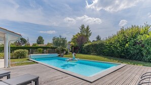 Et rektangulært pool med træterrasse, liggestole og en lille springvand.