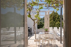 Eine Terrasse mit weißen Stühlen und einem Tisch, eine Pergola mit Weinranken und Blick auf Bäume und Gebäude.