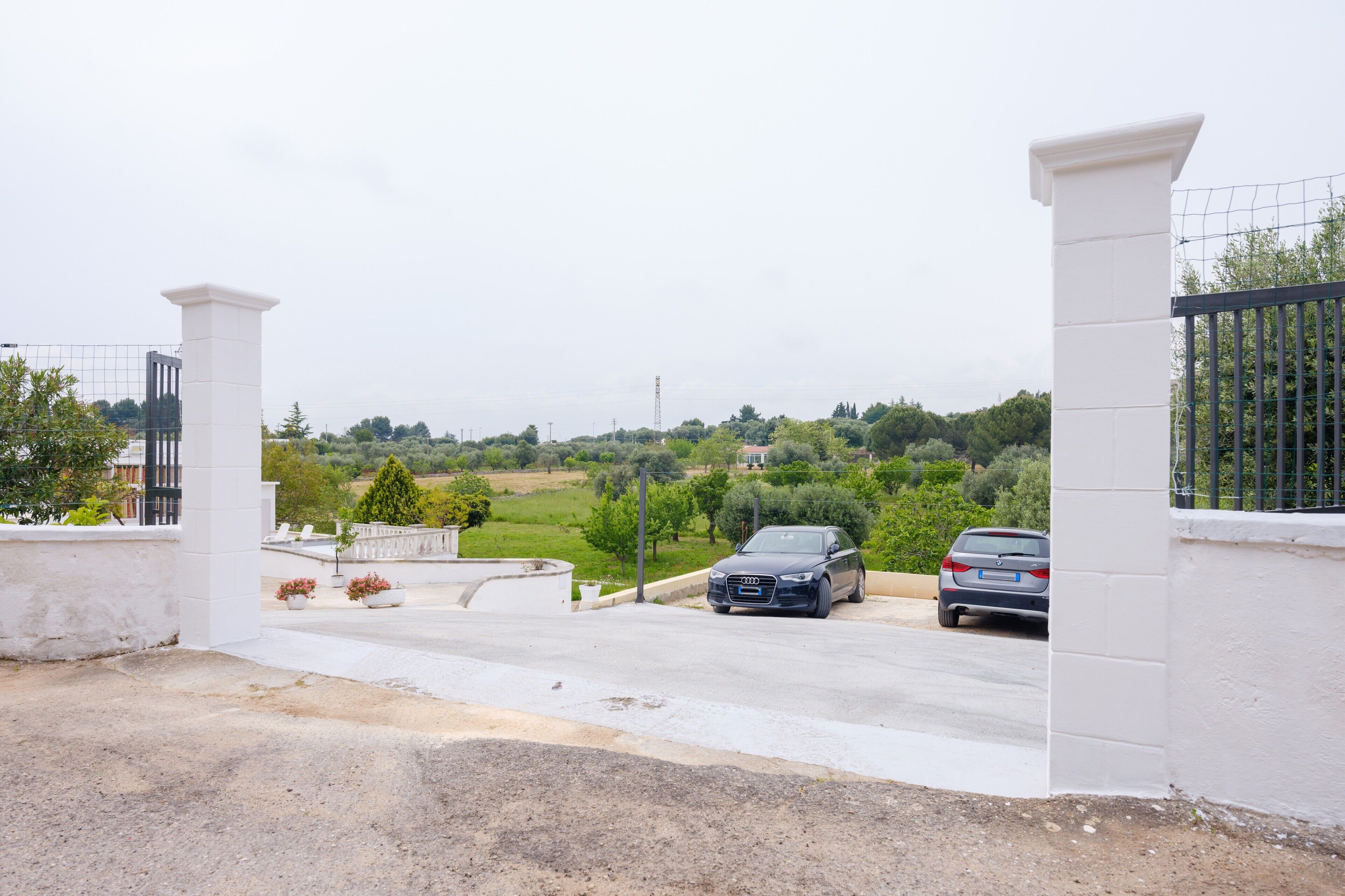 Un vialetto recintato che porta a delle auto parcheggiate, un muro bianco e una vista su un paesaggio verde.