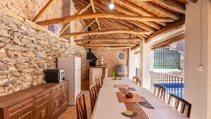 A rustic kitchen with stone walls, wooden beams, a long dining table, and a view of a swimming pool.