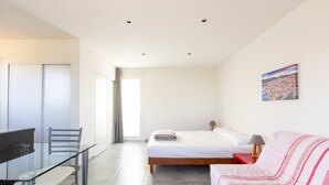 A modern bedroom with a bed, a glass dining table, chairs, a red side table, and a cityscape painting on the wall.