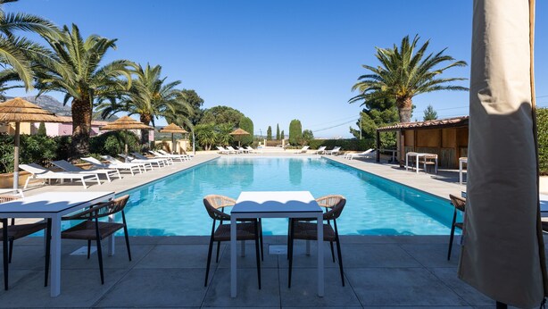 A swimming pool with lounge chairs and tables, surrounded by palm trees and a clear sky.